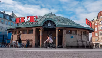 A round, brick building with a green dome roof displays large red BT letters. Two people ride bicycles in front, under a blue sky with scattered clouds. Surrounding the building are older-style structures with ornate facades.