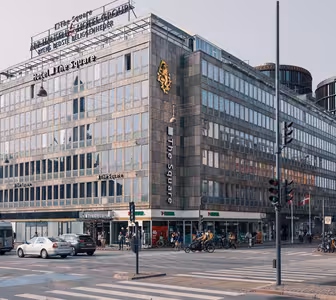 A bustling city street with a large gray building labeled Hotel The Square at the corner. Cyclists and pedestrians move past, and various other buildings are visible. The scene has traffic lights and crosswalks under an overcast sky.