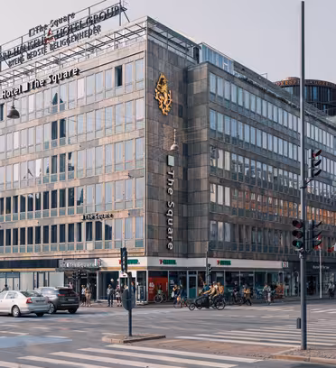 A bustling city street with a large gray building labeled Hotel The Square at the corner. Cyclists and pedestrians move past, and various other buildings are visible. The scene has traffic lights and crosswalks under an overcast sky.