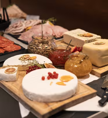 A table with an array of cheeses, cured meats, nuts, and jams displayed. Round cheeses are garnished with dried citrus, while jars contain nuts and spreads. There are cutting boards, knives, and stacks of crackers on the side.