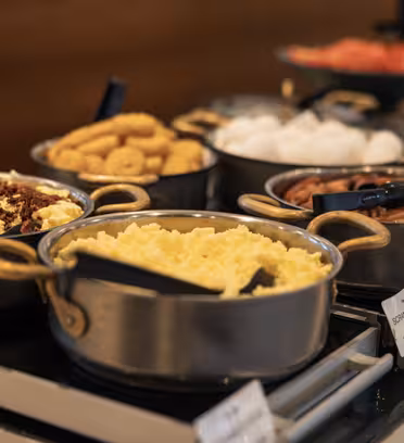A buffet spread featuring various dishes in metal pots. Visible items include scrambled eggs, sausages, and donuts. Utensils are placed in each pot for serving. A wooden backdrop and a plate of sliced fruits are in the background.