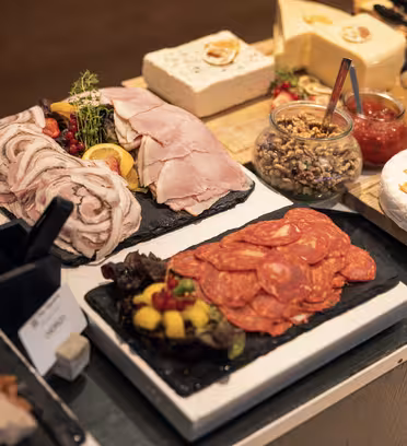 A variety of cold cuts and cheeses are displayed on black platters. Items include sliced ham, salami, and pâté, alongside jars of nuts, fruit garnishes, and blocks of cheese. Small signs label each selection.
