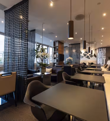 A modern lounge area with black tables and chairs, beige and gray cushioned seating, potted plants, and large windows. A woven partition separates spaces, and a wall features the text CAN RESIST ANYTHING BUT in a stylish design.