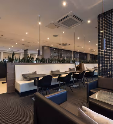 Modern restaurant interior with black and white decor. Booths with white seats and black chairs line one wall, divided by a plant partition. Cozy seating with a small glass table and patterned wall on the right. Ceiling lights add warmth.