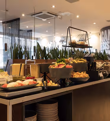 A buffet setup featuring a variety of foods, including bread, pastries, fresh fruits, and parfaits. The display is on a dark wooden counter with a modern interior and plants in the background. Bright lighting highlights the assortment.