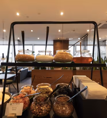 A restaurant buffet setup featuring a two-tiered stand. The top shelf holds jars of condiments with spoons. Below are containers with various toppings and ingredients. The background shows a dining area with modern decor and indoor plants.