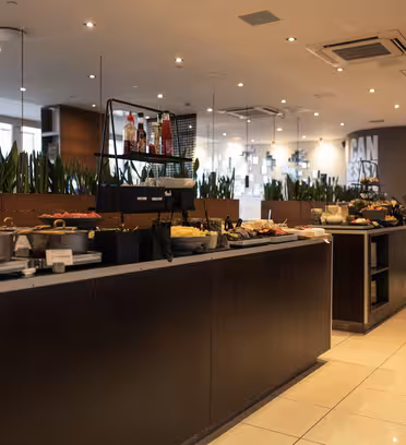 A spacious buffet setup in a modern restaurant. Long tables display various dishes, condiments, and desserts. Green plants separate sections, and overhead lights illuminate the area. The ambiance is clean and inviting with mirrors and a tiled floor.