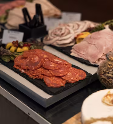 A selection of deli meats including sliced chorizo, ham, and turkey is artfully arranged on dark platters. A jar of walnuts, a cheese wheel, and small bowls of fruit garnish the display on a wooden countertop.