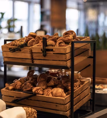 Wooden trays filled with a variety of croissants and pastries are stacked on a metal rack. The setting appears to be a breakfast buffet with a cozy, well-lit atmosphere.