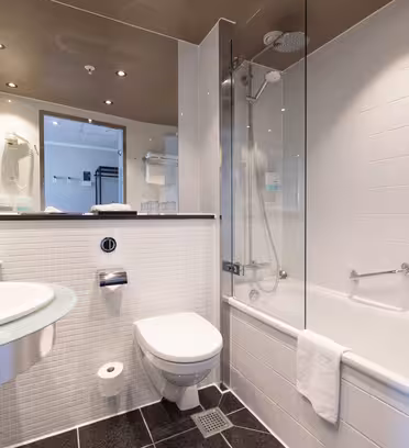Modern bathroom with white tiles, a glass sink, wall-mounted toilet, and a bathtub with a glass shower screen. A towel hangs on the tub, and large mirrors reflect the room’s features.