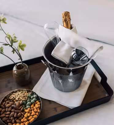 A tray on a bed holds a small vase with green flowers, a bowl of assorted nuts, and a metal ice bucket with a bottle of champagne and two glasses wrapped in a white napkin.