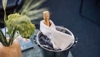 A bottle of champagne wrapped in a white napkin sits in an ice bucket with two glasses. Nearby, a vase with green flowers decorates a table. The setting appears elegant and ready for celebration.
