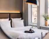 A neatly made bed with white linens and brown headboards is reflected in a mirror. A tray of snacks and drinks sits on the bed, and a vase of green plants sits on the windowsill next to large windows with gray curtains.