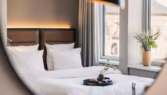 A neatly made bed with white linens and brown headboards is reflected in a mirror. A tray of snacks and drinks sits on the bed, and a vase of green plants sits on the windowsill next to large windows with gray curtains.
