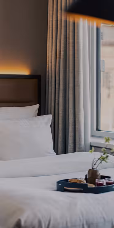 A cozy hotel room with two neatly made beds, white bedding, a tray with breakfast items, large windows with light gray curtains, and a vase with flowers on a windowsill.