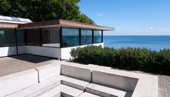 Modern building with large windows overlooking a blue sea and clear sky. Surrounded by lush greenery, the structure features a flat roof and concrete elements in the foreground.