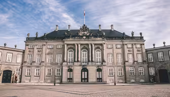 A grand historic building with ornate architecture, featuring tall arched windows, a slate roof, and decorative statues along the rooftop. The front courtyard is paved with cobblestones under a partly cloudy sky.
