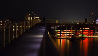 A nighttime scene with a crescent moon in the sky. A long, lit-up boardwalk leads to a pier where brightly colored kayaks are stacked. Buildings are visible in the background, reflecting lights on the water.