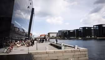 Modern waterfront area with people sitting on chairs by a glass building reflecting the sky. More office buildings in the background across the water. A pink bike is parked nearby, and the sky is partly cloudy.
