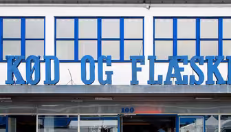 People relax on deck chairs outside a building with blue-framed windows and a sign that reads Kød og Flæsk. A sculpture of a cow is displayed above the entrance. The scene is lively and social, with a casual outdoor atmosphere.