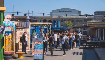 A lively outdoor market scene with people gathered around food stalls. Colorful signs and string lights add vibrancy. In the background, a building displays Reffen and Refshaleøen in large letters.