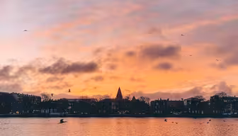 A serene sunset over a calm lake with the silhouette of a city skyline featuring a prominent spire in the distance. The sky is painted in shades of pink, orange, and purple, with scattered clouds and birds flying above the water.