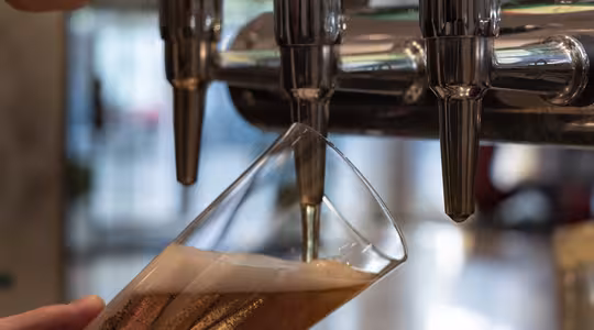 A close-up of a hand tilting a glass under a beer tap as amber-colored beer is being poured, creating a foamy head. The background is blurred, highlighting the focus on the filling process.