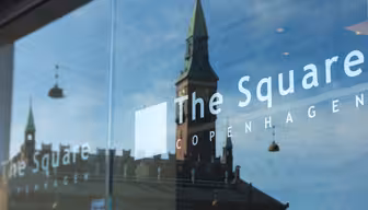 The photo shows a glass window reflecting a historic building with a green-roofed tower and streetlights against a blue sky. The Square and Copenhagen are written on the glass.