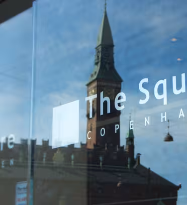 The photo shows a glass window reflecting a historic building with a green-roofed tower and streetlights against a blue sky. The Square and Copenhagen are written on the glass.