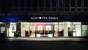Night view of a hotel entrance with a sign that reads Hotel The Square. The facade is illuminated, showcasing large glass windows and a modern interior with seating visible inside.