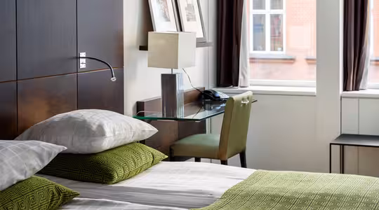 Modern hotel room with a neatly made bed featuring green and white linens. A glass desk with a lamp, phone, and chair is by the window. Framed pictures hang above the bed, and large windows overlook a brick building.