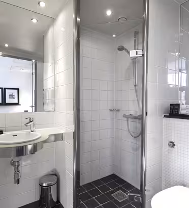 Modern bathroom with a glass-enclosed shower, white tiled walls, and a dark tiled floor. Features a wall-mounted sink, towels on a rack, a hairdryer, and a large mirror. Natural light enters from the window on the far wall.