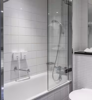 Modern bathroom with white tiled walls and a glass-enclosed bathtub. The showerhead is mounted on the wall, and toiletries are attached inside the shower area. A white toilet and sink are visible, with towels folded on a shelf.