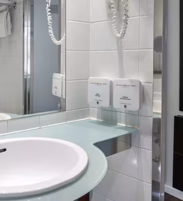 Modern bathroom with a white sink and chrome faucet set in a light green countertop. The wall has white tiles, a mounted hairdryer, and two soap dispensers. A hanging towel is visible on a rack near a glass shower door. A mirror reflects the scene.