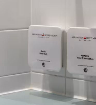 Two wall-mounted dispensers from Arp-Hansen Hotel Group in Copenhagen are attached to a tiled bathroom wall. The left dispenser contains Gentle Hand Wash, and the right contains Hydrating Hand & Body Lotion.