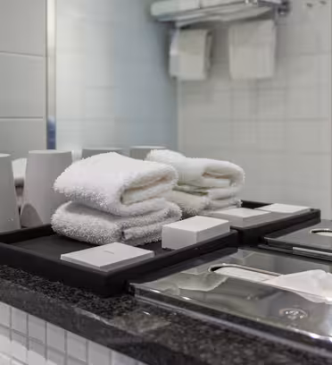 A bathroom counter with folded white towels, two soap trays, tissue boxes, and two cups. A large mirror reflects the neatly arranged items, with a glass door visible in the background.