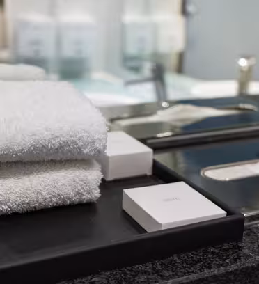 A bathroom countertop with neatly stacked white towels and a bar of soap on a black tray. A tissue box is visible next to them, and a mirror reflects the scene, adding depth to the image.