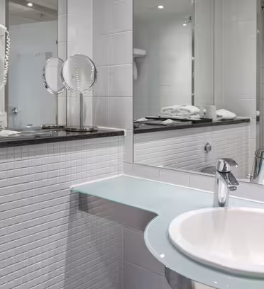Modern hotel bathroom with a white sink, glass countertop, and a wall-mounted mirror. Two hairdryers are mounted on a tiled wall. Towels and toiletries are arranged neatly on a black surface above the sink.