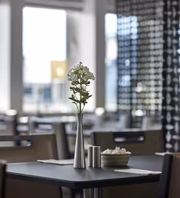 A modern, bright café with a focus on a table setting. A white flower in a metallic vase is placed on the table, along with a small dish and cup. The background shows blurred chairs and decorative wall panels.