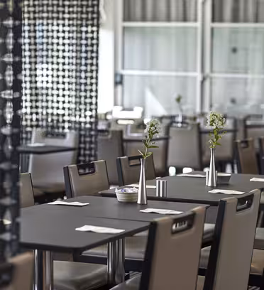Modern dining room with rows of tables and chairs. Each table has a small vase with flowers and neatly placed napkins. The room is partially divided by decorative black screens, adding a stylish touch. Bright natural light filters through large windows.