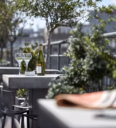 A patio table with two empty wine glasses, a green wine bottle, and a small bowl. Surrounded by greenery and other tables, it overlooks buildings in the distance. The foreground shows a blurred coffee cup and book on another table.