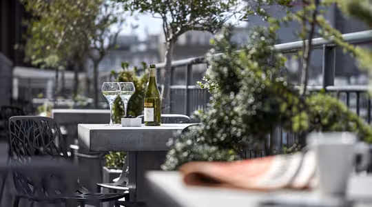 A patio table with two empty wine glasses, a green wine bottle, and a small bowl. Surrounded by greenery and other tables, it overlooks buildings in the distance. The foreground shows a blurred coffee cup and book on another table.