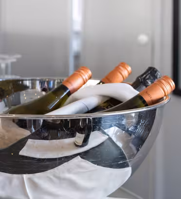 A metallic bowl containing several bottles of wine, each with an orange cap, partially wrapped in a white cloth, sits on a table. A red-capped wine bottle and upside-down wine glasses are nearby, against a backdrop of white and glass surfaces.