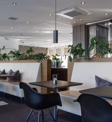 Modern restaurant interior with black chairs and tables, beige upholstered seating, and greenery throughout. Ceiling lights illuminate the space, creating a clean and contemporary atmosphere.