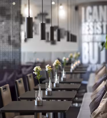 Modern café with a row of black tables and chairs adorned with small flower vases. Pendant lights hang from the ceiling. In the background, a wall partly displays the text, I can resist everything but.... Indoor plants add greenery.
