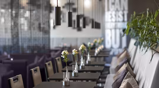 Modern café with a row of black tables and chairs adorned with small flower vases. Pendant lights hang from the ceiling. In the background, a wall partly displays the text, I can resist everything but.... Indoor plants add greenery.