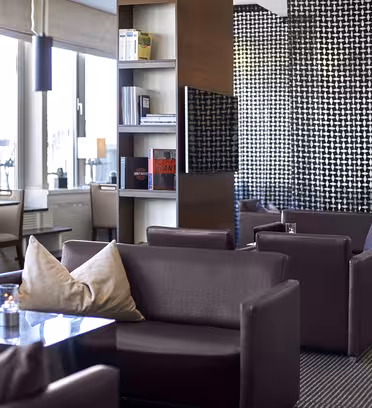 Modern lounge with dark brown armchairs and small tables, each adorned with a candle and cushion. Shelves divide the space, and a large window allows natural light in. Black and white geometric patterns decorate the walls.
