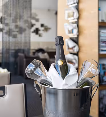 A bottle of champagne is chilled in an ice bucket with two glasses nearby. The scene is set in a modern restaurant with contemporary decor, featuring books and geometric wall art in the background.
