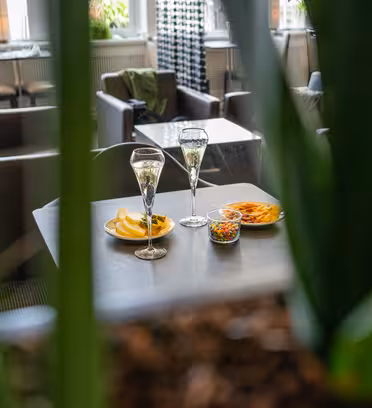 Two glasses of champagne on a table with plates of chips and mixed nuts. Blurred green leaves in the foreground frame the cozy, modern interior with comfortable seating.