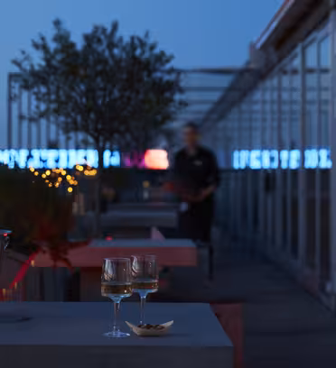 Two glasses of wine sit on a table at a rooftop bar during dusk. A bucket with a bottle is nearby. Twinkling string lights decorate the area, and a blurred figure is in the background. The city skyline is softly illuminated.